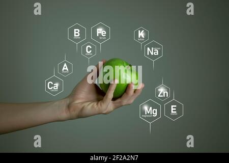 Frau`s Hand hält grünen Apfel, Mikroelement-Symbole in molekularen Sechsecken auf grauem Hintergrund. Vorlage für ein gesundes Lebensmittel- und Lifestyle-Konzept für das Produkt Stockfoto